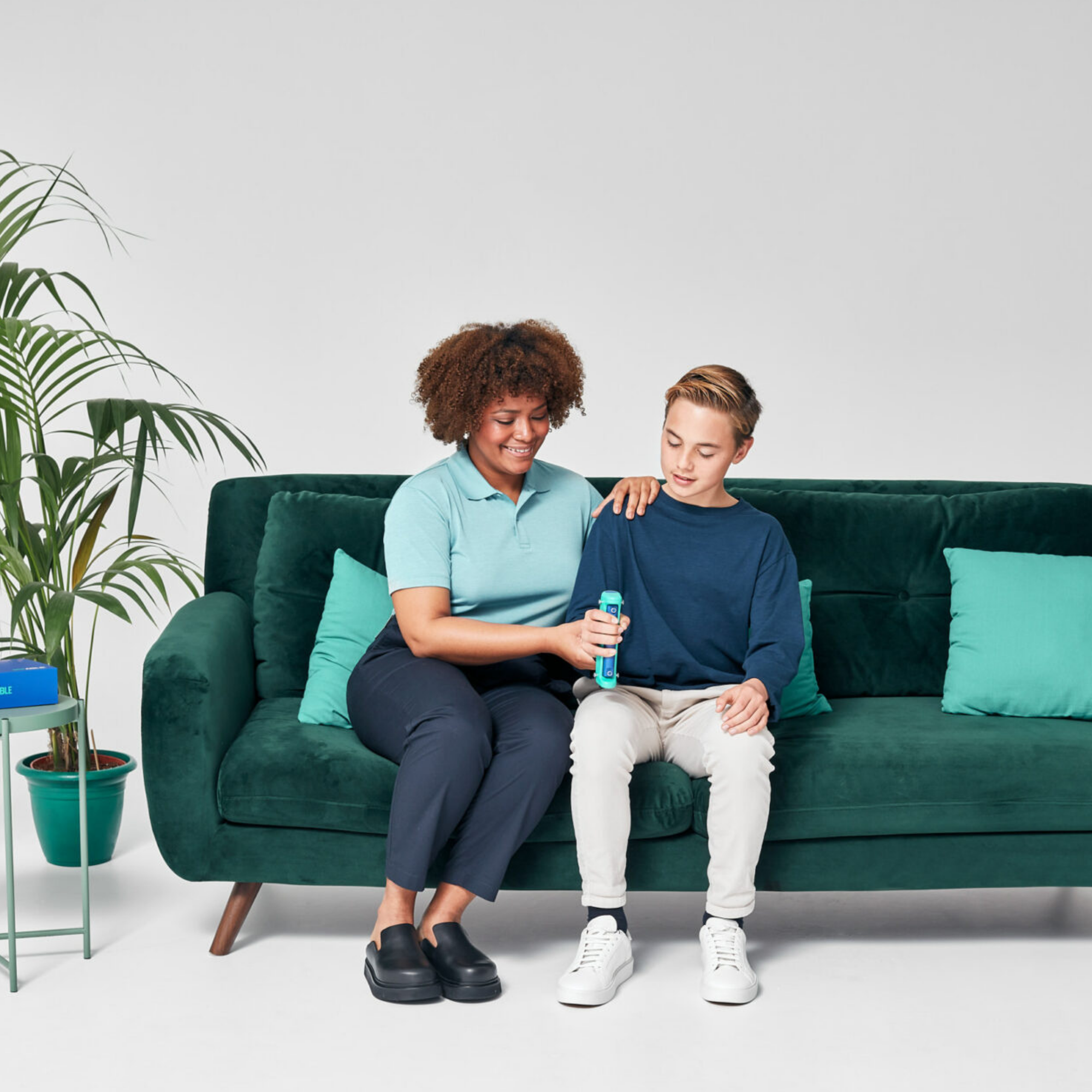A woman and a boy sitting on a dark green velvet sofa, holding a turquoise bottle together in a bright, modern room with a potted plant on the left.