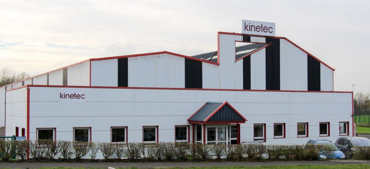A large white industrial building with black and red vertical stripe accents, featuring the 'kinetec' logo on the roof and facade, set against an overcast sky.