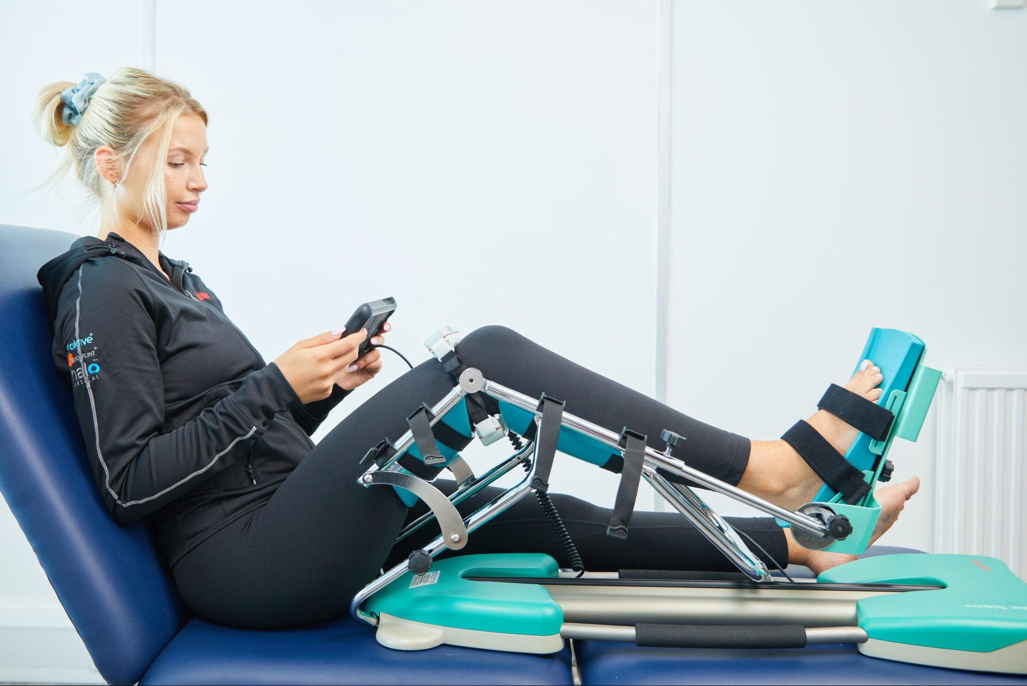 A woman reclining on a blue treatment table using a leg exercise device with teal padding and black straps, while holding a smartphone in a bright indoor room.