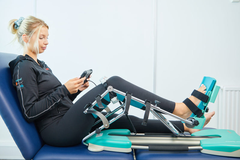A woman reclining on a blue treatment table using a leg exercise device with teal padding and black straps, while holding a smartphone in a bright indoor room.