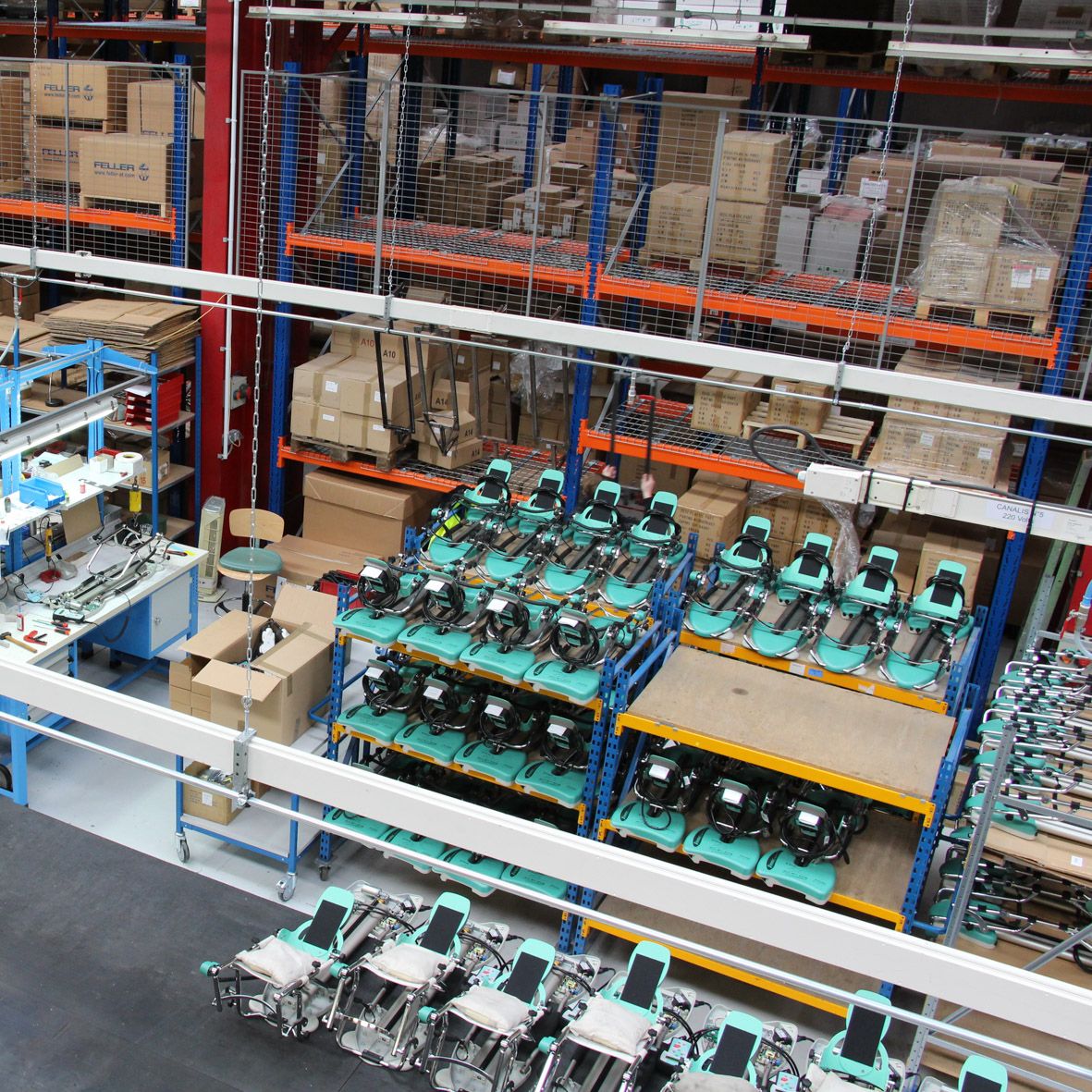 A high-angle view of a warehouse assembly line with multiple teal and black power tools organized on blue shelving units, surrounded by cardboard boxes and industrial storage racks.
