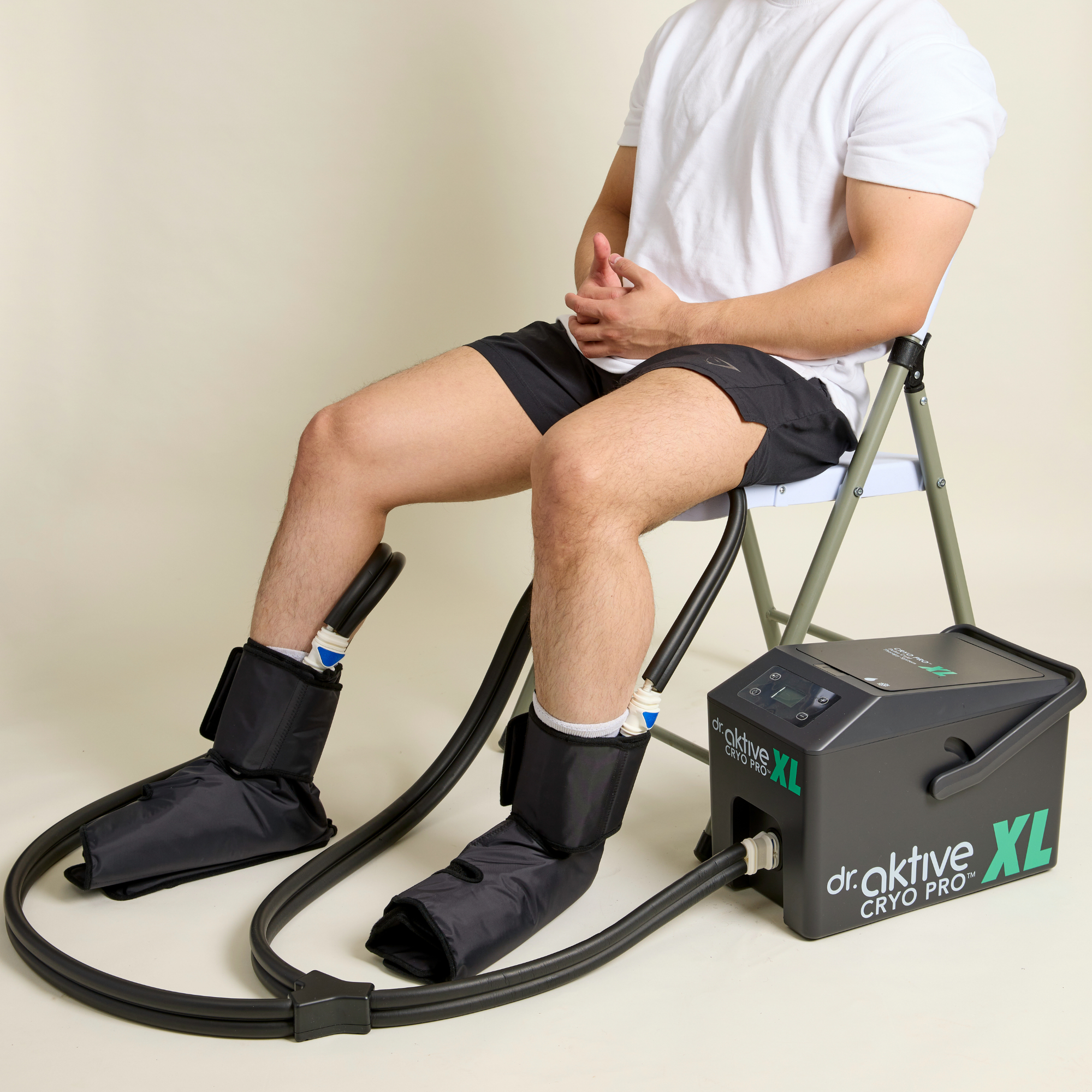 A man sits on a chair using a dr. aktive CRYO PRO XL cryotherapy device with black compression cuffs on both legs, shown against a plain beige studio background.