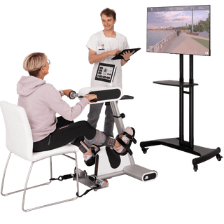 A seated person uses a white and gray leg exercise machine with a hand-held resistance unit, while a standing trainer holds a tablet. A large screen on a black stand displays a virtual outdoor walkway.