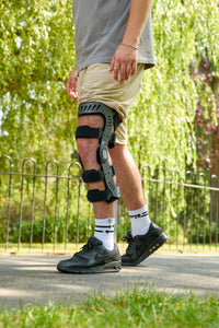 A person outdoors wearing a black and gray knee immobilizer brace on their right leg, paired with khaki shorts, white socks, and black sneakers, with a park background.