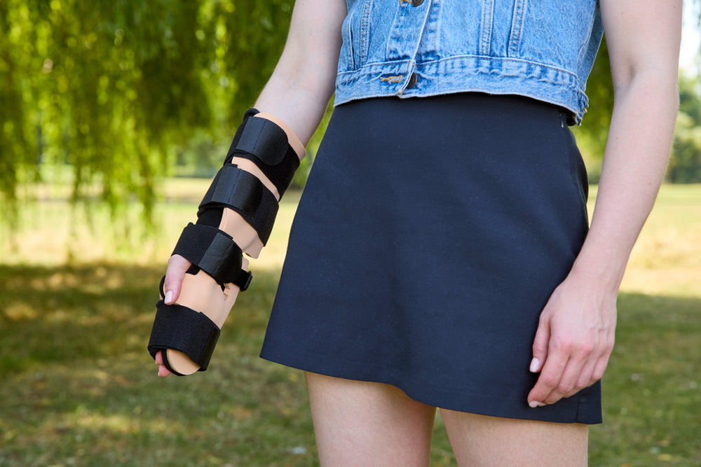 A person outdoors wearing a black multi-band forearm and wrist brace on one arm, paired with a denim top and dark skirt, with blurred green trees in the background.