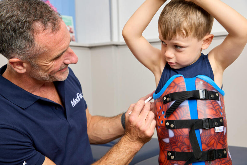 A young boy wearing a red and blue back support brace with black straps, having his straps adjusted by a male healthcare provider in a clinical setting.