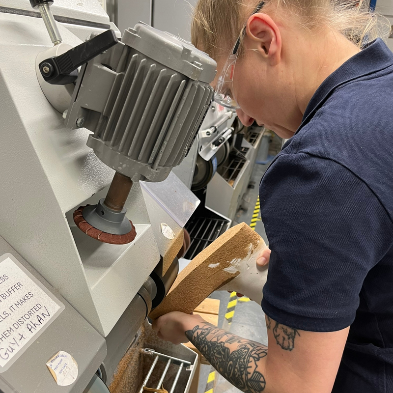 A person in a navy blue polo shirt and safety glasses uses a handheld tool to shape a cork block against a large industrial grinding machine in a workshop setting.