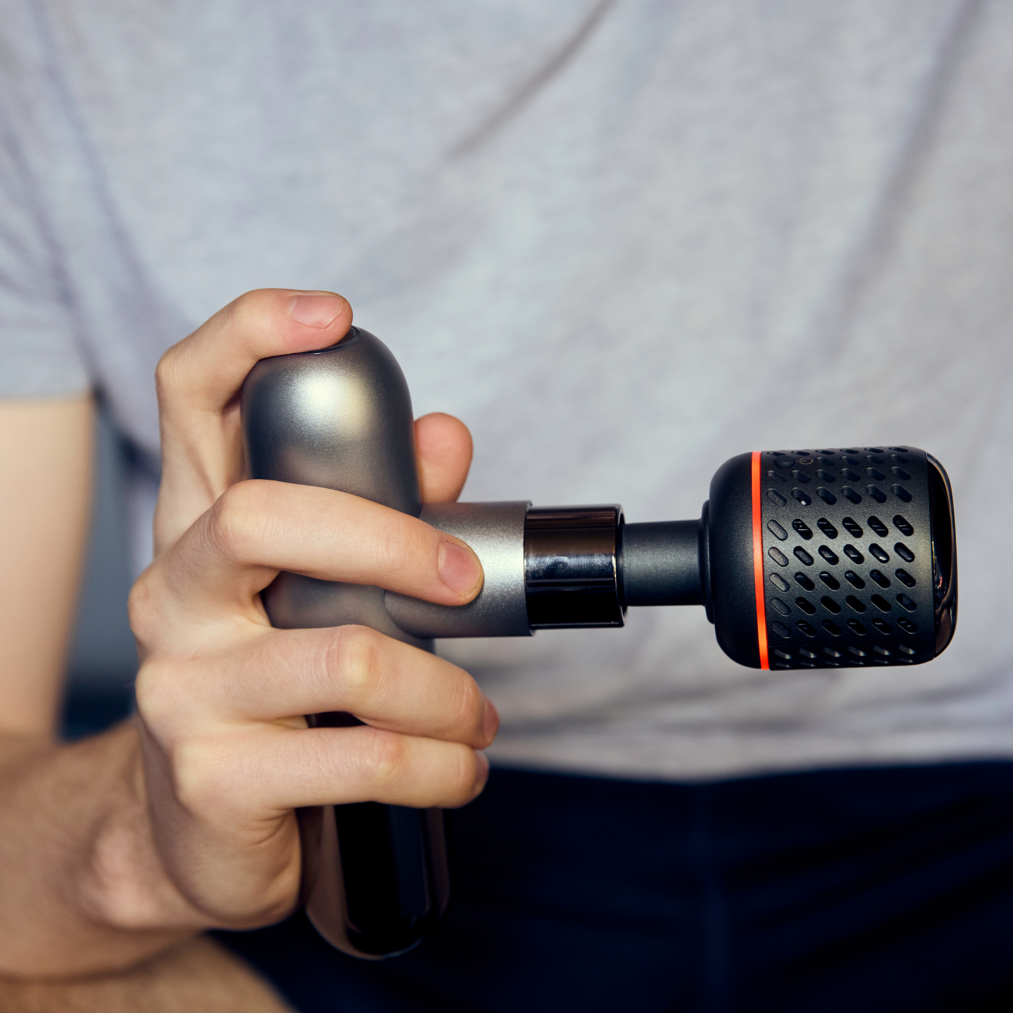 A close-up of a person holding a handheld massage gun with a textured black head and a metallic dark gray body. The person is wearing a light gray t-shirt and dark pants, and the background is a soft, out-of-focus neutral gray.
