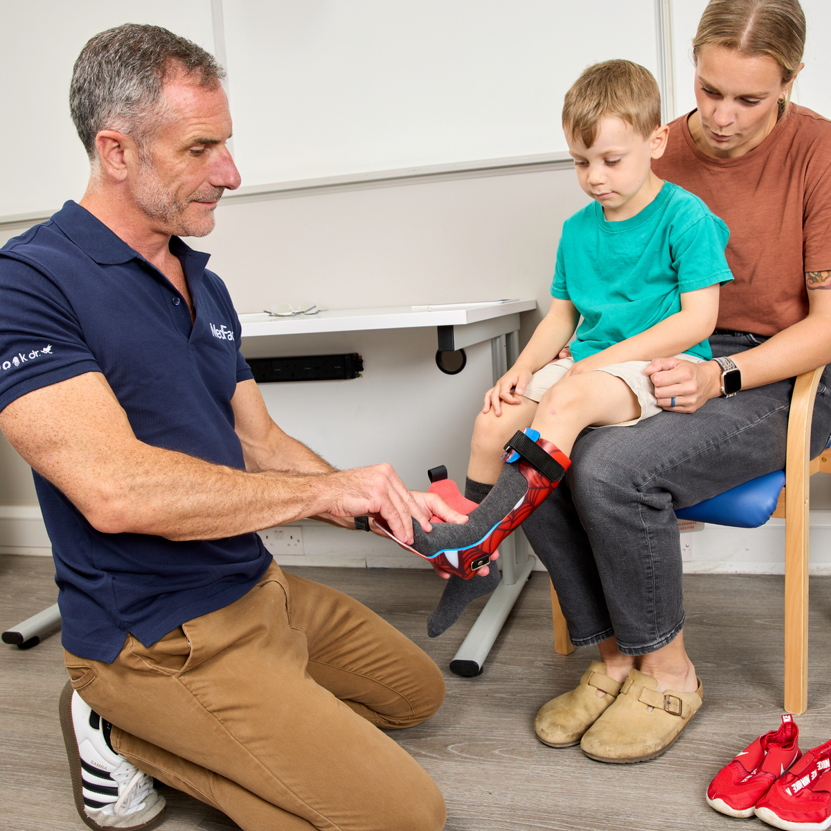 A man kneels on the floor while a child sits on a woman's lap, receiving help to put on a red and black orthopedic shoe. The child is wearing a turquoise t-shirt and beige slip-on shoes, and the man wears a navy blue polo shirt.